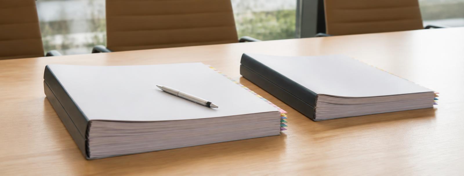 Two thick bound submission packs with tabbed dividers on a boardroom table, a pen resting on top